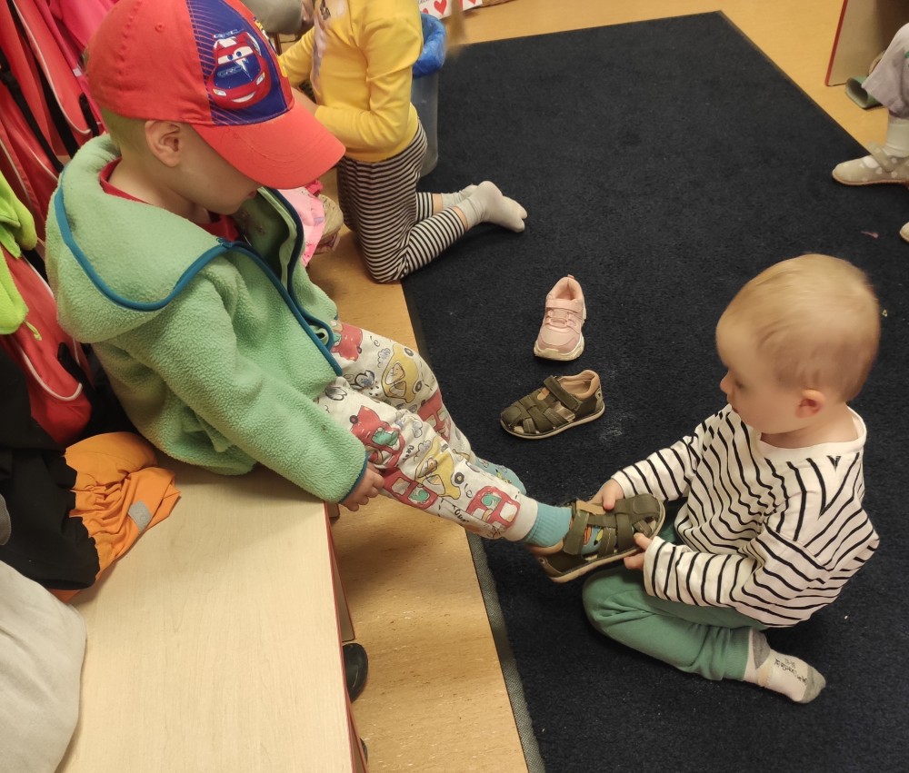 Trzyletnie dziecko pomaga rówieśnikowi zakładać sandały,
przykład wzajemnej pomocy wśród przedszkolaków