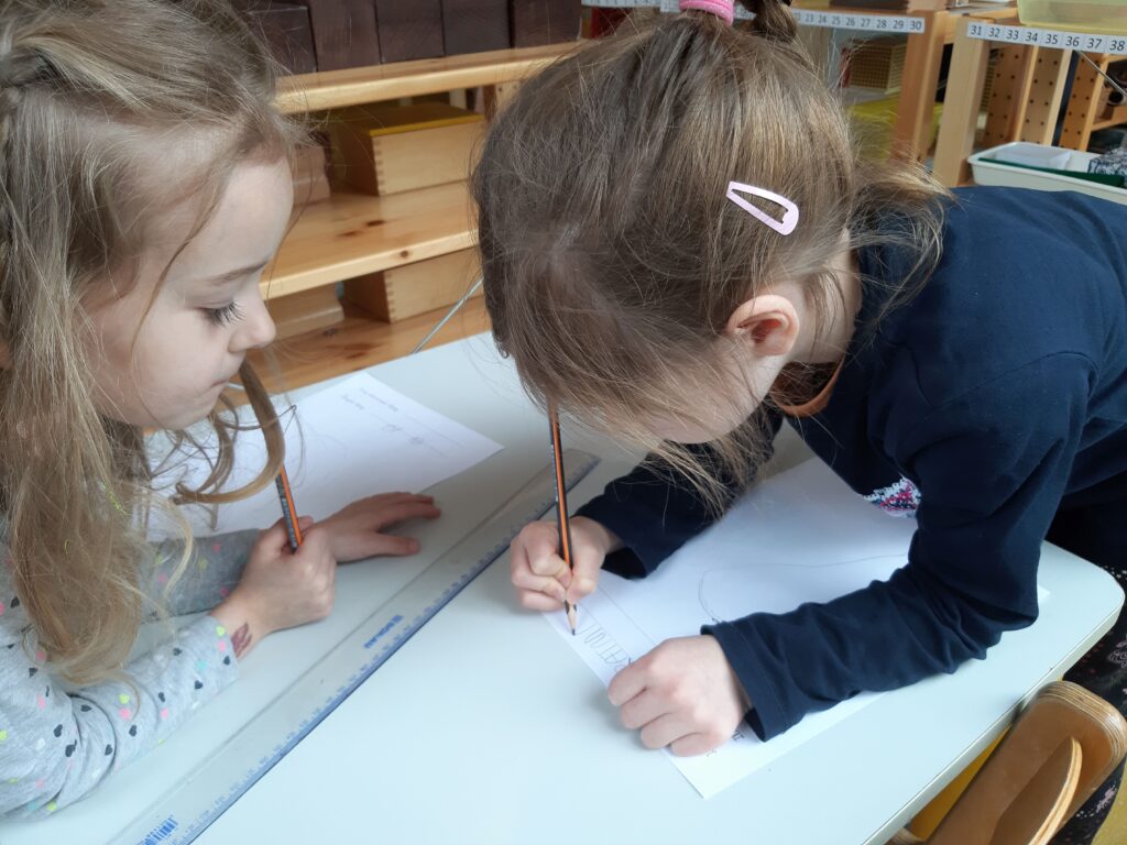 Starsza dziewczynka uczy młodszą pisania; dziecko trzyma ołówek i ćwiczy litery