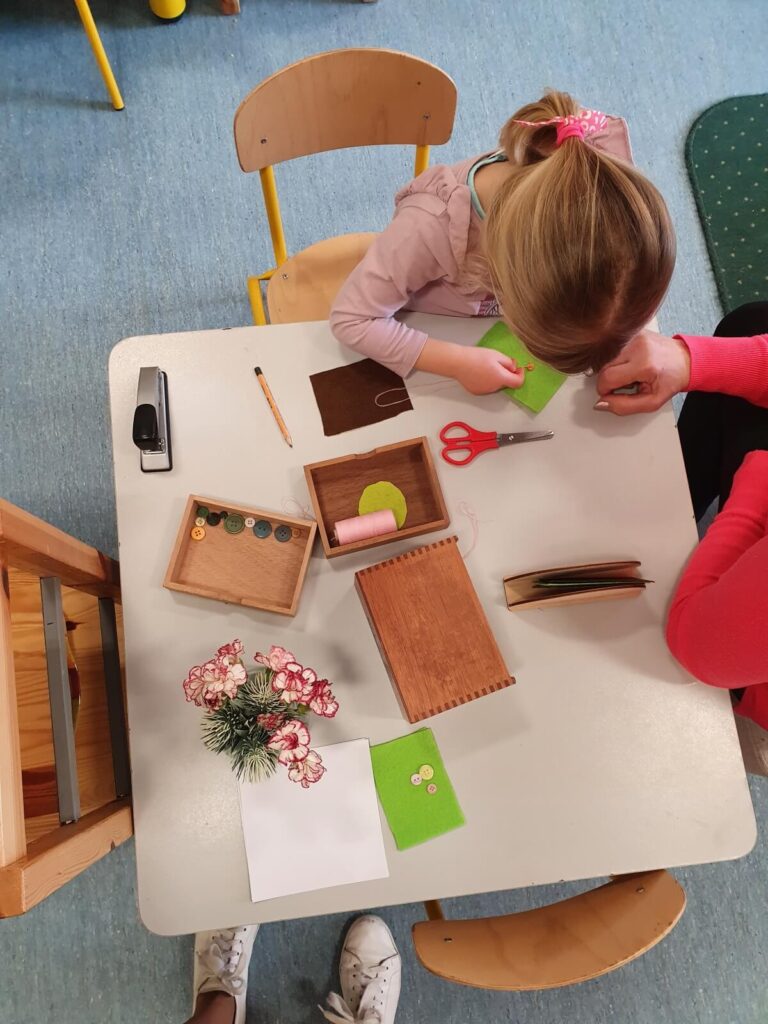 Sześcioletnia Marysia przyszywa guzik do kawałka tkaniny, wciągając nitkę igłą z dużym uszkiem.